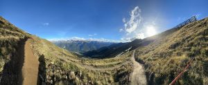 Ein Panoramablick auf eine Berglandschaft mit grasbewachsenen Hügeln, gewundenen Mountainbike-Strecken und fernen Bergen unter einem klaren blauen Himmel. Die Sonne steht tief und wirft helles Licht und lange Schatten auf die malerischen Strecken.