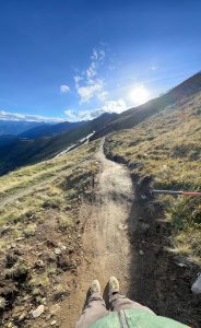 Ein schmaler Feldweg schlängelt sich unter strahlend blauem Himmel an einem grasbewachsenen Berghang entlang, während die Sonne über den fernen Gipfeln scheint. Unten ist die Spitze eines Wanderschuhs zu sehen, was an Mountainbike-Abenteuer auf diesem malerischen Weg erinnert.