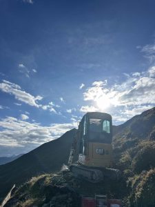 Ein gelber CAT-Bagger steht an einem felsigen Berghang und formt unter einem hellen, teilweise bewölkten Himmel einen neuen Mountainbike-Trail. Die Sonne scheint hinter der Maschine, und grasbewachsene Hügel und Berge umgeben die malerische Mountainbike-Landschaft.