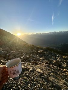 Eine Hand hält einen weißen Becher mit der Aufschrift „Bikepark“ und blickt auf einen felsigen Bergpfad bei Sonnenaufgang oder Sonnenuntergang. Die Sonne steht nahe dem Horizont und wirft goldenes Licht auf die fernen Berge und das Tal darunter.