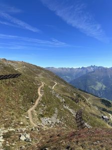 Ein kurvenreicher Bergpfad, ideal für Mountainbiker, mit hölzernen Schneezäunen auf einem Grashang unter klarem blauen Himmel und zarten Wolken. Ferne Berge und Täler bereichern die malerische Strecke.