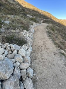 Ein schmaler Feldweg schlängelt sich durch felsiges Gelände bergauf, auf der einen Seite von einer niedrigen Steinmauer und auf beiden Seiten von trockenem Gras und Sträuchern begrenzt, was ihn zu einer idealen Mountainbike-Strecke unter einem klaren, blauen Himmel macht.