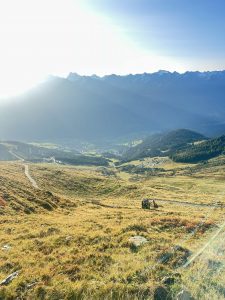 Sonnenlicht scheint über ein Gebirgstal mit grünen Feldern und Wäldern. Ein kleiner Bagger steht auf dem grasbewachsenen Hang neben einem gewundenen Mountainbike-Trail, der unter strahlend blauem Himmel zu weit entfernten Dörfern führt.