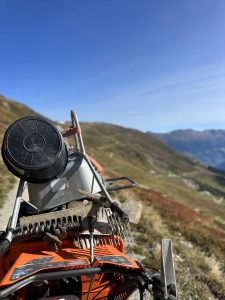 Nahaufnahme von Gartengeräten, darunter ein Rechen und ein Eimer, auf einem Wagen oder einer Maschine neben einem Mountainbike-Trail in einer grasbewachsenen Berglandschaft unter klarem blauen Himmel. Im Hintergrund sind sanfte Hügel und Berge in der Ferne zu sehen.