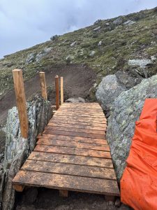Eine kleine Holzbrücke mit einfachem Geländer überspannt felsiges Gelände auf einem Mountainbike-Trail. Der Boden ist grasbewachsen, eine orangefarbene Plane bedeckt einen Teil des Felsens rechts, im Hintergrund ist ein bewölkter Himmel zu sehen.