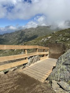 Eine hölzerne Fußgängerbrücke mit Geländer überquert felsiges Gelände auf einem Bergpfad, ideal für Mountainbike-Bewegung, umgeben von grünen Hängen und teilweise bewölktem Himmel mit fernen Gipfeln im Hintergrund.