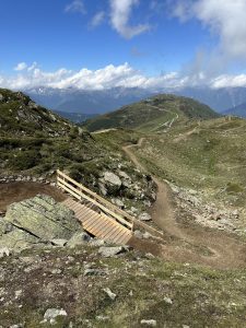 Ein gewundener Feldweg, der sich perfekt für ein Mountainbike-Abenteuer eignet, führt über eine kleine Holzbrücke zwischen felsigen, grasbewachsenen Hügeln mit Bergen und einem teilweise bewölkten blauen Himmel im Hintergrund.