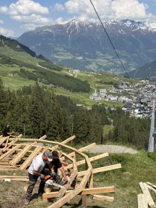 Drei Personen bauen auf einem grasbewachsenen Hügel eine Holzkonstruktion auf, die perfekt für Mountainbike-Enthusiasten ist und unter einem teilweise bewölkten Himmel eine malerische Aussicht auf eine Stadt, Kiefern und schneebedeckte Berge im Hintergrund bietet.