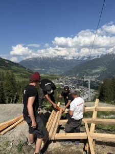 Vier Männer arbeiten zusammen und bauen an einem sonnigen Tag Holzbretter an einem Hang, um einen Pfad mit malerischer Aussicht auf Berge, ein Tal und eine darunterliegende Stadt zu bauen.