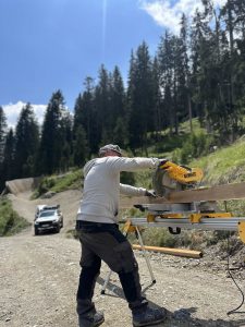 Eine Person in Arbeitskleidung verwendet eine DeWalt-Gehrungssäge, um Holz auf einem Ständer im Freien zu schneiden. Ein Mountainbike-Track schlängelt sich an einem bewaldeten Hang entlang und im Hintergrund ist unter einem teilweise bewölkten Himmel ein weißer Lastwagen zu sehen.