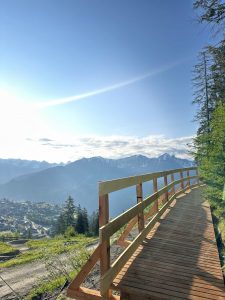 Ein Holzweg schlängelt sich an einem Berghang entlang, von dem aus man ein Tal überblicken kann, in dem sich eine Stadt befindet. In der Ferne erheben sich hohe Berge mit schneebedeckten Gipfeln unter einem klaren, sonnigen Himmel. Bäume säumen den Weg rechts.