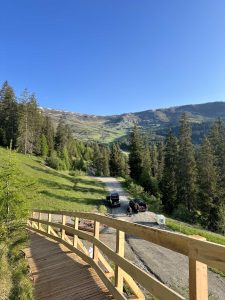 Ein Holzgeländer säumt einen Weg mit Blick auf einen Schotterweg, unter dem zwei Autos parken. Hohe Kiefern umgeben das Gebiet, im Hintergrund erheben sich grüne Berge unter einem klaren blauen Himmel. Ideal für ein Mountainbike-Abenteuer.