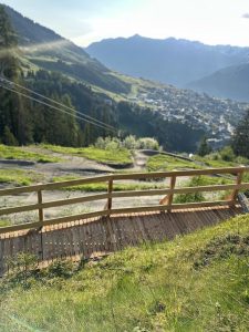Eine hölzerne Fußgängerbrücke führt über einen grasbewachsenen Berghang. Darunter verläuft ein gewundener Mountainbike-Pfad, immergrüne Bäume und in einem Tal liegt eine Stadt – alles umgeben von fernen, dunstigen Bergen unter einem klaren Himmel.
