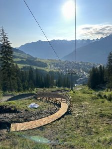 Ein Holzpfad schlängelt sich unter einer Seilbahnlinie einen grasbewachsenen Hügel hinunter, mit einem sonnigen Himmel, Kiefern und einem zwischen Bergen eingebetteten Dorf im Hintergrund.