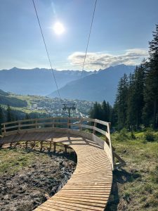 Ein geschwungener Holzsteg bietet Aussicht auf ein grünes Tal, eine entfernte Stadt und schneebedeckte Berge unter einem sonnigen blauen Himmel. Über ihm verläuft ein Skiliftkabel, und auf der rechten Seite sind hohe Kiefern zu sehen.
