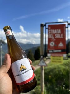 Eine Hand hält eine Bierflasche mit der Aufschrift „Mountain Bike Movement“ vor einem Mountainbike-Trail-Schild mit der Aufschrift „Strada del Sole“, mit blauem Himmel und grünen Hügeln im Hintergrund.