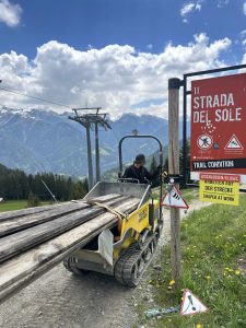 Ein Arbeiter steuert ein kleines Fahrzeug, das Holzbretter transportiert, in der Nähe eines Skipistenschilds mit der Aufschrift „Strada del Sole“. Im Hintergrund sind Berge, Bäume und ein Skilift unter einem bewölkten Himmel zu sehen – die perfekte Kulisse für ein Mountainbike-Abenteuer.