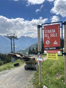 Ein Bergpfad wird gewartet. Ein Karren transportiert Holzbretter auf dem Feldweg. Ein rotes Schild mit der Aufschrift „Strada del Sole“ weist darauf hin, dass der Weg wegen Bauarbeiten gesperrt ist. Berge und Wolken bilden eine malerische Kulisse – normalerweise ein beliebtes Ziel für Mountainbike-Abenteuer.