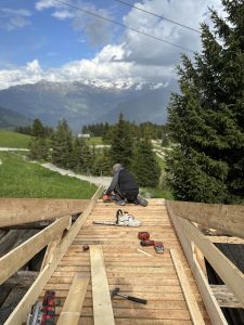 Eine Person baut im Freien, umgeben von grünem Gras, Kiefern und Bergen, unter einem teilweise bewölkten Himmel einen hölzernen Mountainbike-Trail. Verschiedene Werkzeuge und Materialien liegen verstreut auf der Holzoberfläche.