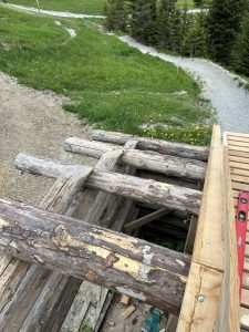 Blick von oben auf eine im Bau befindliche Holzbrücke mit großen Baumstämmen als Stützbalken. Die Konstruktion ist Teil einer Mountainbike-Strecke an einem grasbewachsenen Hang. Im Hintergrund sind ein Feldweg und Bäume zu sehen.