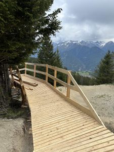Ein Holzsteg mit einer Bank schlängelt sich durch einen Bergwald, perfekt für Mountainbike-Fans, und bietet Aussicht auf ein Tal mit grünen Feldern und in der Ferne schneebedeckte Gipfel unter einem bewölkten Himmel.