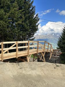 Eine hölzerne Fußgängerbrücke mit Geländer überspannt einen Feldweg in einer bergigen Gegend, umgeben von hohen Kiefern. Schneebedeckte Berge und ein teilweise bewölkter Himmel bilden die Kulisse und laden jeden Mountainbike-Enthusiasten zu Abenteuern ein.