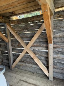 Innenansicht einer Holzschuppenwand aus horizontalen dunklen Baumstämmen, verstärkt durch zwei hellere diagonale Balken, die eine X-Form bilden – eine Erinnerung an die robusten Stützstrukturen entlang eines schroffen Mountainbike-Trails.