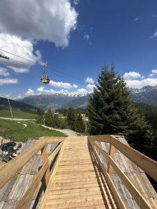 Eine Holzrampe führt hinunter zu grasbewachsenen Hügeln und Kiefern und markiert den Beginn eines Mountainbike-Trails. Eine gelbe Seilbahn hängt an einem Drahtseil, im Hintergrund schneebedeckte Berge und ein teilweise bewölkter blauer Himmel.