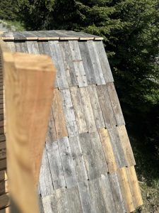 Eine Nahaufnahme eines schrägen Holzdachs aus verwitterten, bunten Brettern, mit einem verschwommenen Holzgeländer im Vordergrund und grünen Bäumen, die im Hintergrund einen Mountainbike-Pfad säumen.