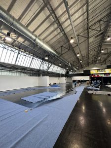 Große Halle mit hohen Decken und Schächten, in der mehrere Personen einen blauen Teppich auf dunklem Boden verlegen. Die Baumaterialien lassen darauf schließen, dass in diesem großzügigen Bereich ein Mountainbike- oder MTB-Event stattfinden wird.