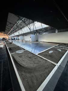 Großer Innenraum mit hohen Decken und Oberlichtern, mit rechteckigen Abschnitten aus dunklem Boden, die einer MTB-Strecke ähneln, und einem zentralen Bereich, der mit reflektierendem Kunststoff bedeckt ist und möglicherweise Teil einer Ausstellung oder eines Bauprojekts ist.