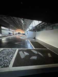 Ein großer Innenraum mit hohen Decken und Oberlichtern zeigt eine teilweise angelegte Mountainbike-Strecke, die von Abschnitten aus Steinen, Kies und Erde gesäumt ist. Im Hintergrund sind weiße Trennwände und Baumaterialien zu sehen.