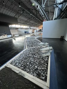 Ein moderner Innenraum mit einem breiten, pfadähnlichen Weg aus Kies und Steinen, eingefasst von Holzrahmen, unter einer hohen Metalldecke mit sichtbaren Balken und Lichtern. Im Hintergrund sind Baumaterialien sichtbar. Perfekt für Mountainbike-Enthusiasten.