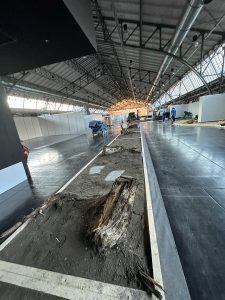 Ein großer Innenraum mit hohen Decken bietet einen langen, unbefestigten Mountainbike-Pfad, der von Holzplanken gesäumt ist. Baumstämme und Schutt säumen die Strecke, während im Hintergrund Menschen damit beschäftigt sind, die Ausrüstung für das Mountainbikemovement-Event aufzubauen.