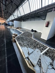 Ein Ausstellungsraum im Innenbereich weist einen langen, gewundenen Pfad aus Felsen, Steinen und großen flachen Platten auf, der von Holzbrettern begrenzt wird. Hohe Decken und weiße Trennwände sind zu sehen, ebenso wie ein an einer Wand montierter Feuerlöscher.