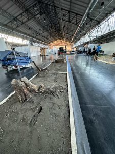 Ein großer Innenraum mit Industriedecken bietet neben einem glatten schwarzen Gehweg einen Ausstellungsbereich mit Erde, Baumwurzeln und Baumstümpfen – eine Inspiration für Mountainbike-Bewegung und MTB-Trails. Im Hintergrund stehen mehrere Personen neben abgedeckten Objekten und Motorrädern.
