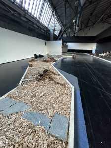 Ein moderner Innenraum mit hohen Decken verfügt über einen gewundenen Holzpfad voller Steine und Holzspäne, der mit blauem Band eingefasst und von einem dunklen, polierten Boden und weißen Wänden umgeben ist.