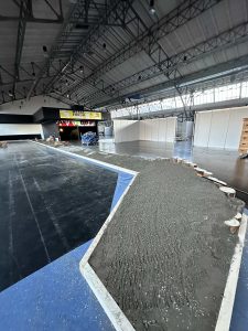 Ein großer Innenraum mit hohen Decken, in dem ein teilweise fertiggestellter, von Baumstämmen gesäumter Mountainbike-Pfad (MTB) aus Beton neben einem schwarz-blauen Streckenboden und weißen Trennwänden im Hintergrund zu sehen ist.