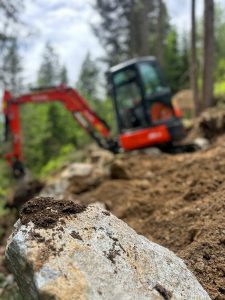 Eine Nahaufnahme eines großen Felsens mit Erde darauf deutet auf eine neue Strecke hin, während ein unscharfer roter Bagger in einem Waldgebiet dahinter arbeitet - ein Hinweis auf den Bau von Mountainbike-Strecken im Freien oder auf Landschaftsbauarbeiten.