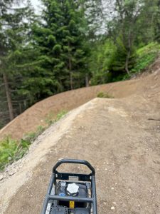 Ein unbefestigter Weg schlängelt sich durch ein bewaldetes Gebiet mit hohen immergrünen Bäumen. Im Vordergrund sind Baumaschinen zu sehen, was darauf hindeutet, dass die Mountainbike-Strecke gerade gebaut oder gewartet wird.
