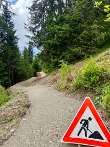 Ein Baustellenwarnschild steht neben einem gewundenen Schotterweg durch einen dichten grünen Wald und weist Wanderer auf mögliche Arbeiten an der Strecke hin.