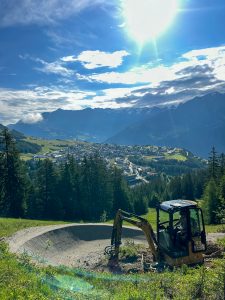 Ein kleiner Bagger arbeitet auf einer Mountainbike-Strecke entlang eines grasbewachsenen Abhangs, mit einer malerischen Talstadt und bewaldeten Hügeln im Hintergrund unter einem hellen, sonnigen Himmel, der von vereinzelten Wolken durchzogen ist.