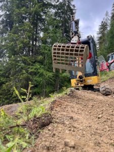 Ein gelber Bagger mit einer Gitterschaufel gräbt oder bewegt Erde auf einer abschüssigen, unbefestigten Fläche in einem Wald mit hohen grünen Bäumen - das perfekte Terrain für Mountainbike-Enthusiasten und die wachsende Mountainbikemovement.