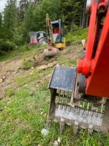 Zwei kleine Bagger mit angehängten Harken arbeiten an der Räumung eines grasbewachsenen, abschüssigen Weges in der Nähe eines Waldes und unterstützen die Mountainbikemobilität. Die Schaufel der näheren Maschine ist scharf abgebildet, während die Maschinen im Hintergrund leicht verschwommen erscheinen.