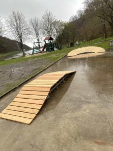 Ein nasser Outdoor-Park verfügt über zwei kleine hölzerne MTB-Rampen auf einer Betonoberfläche, mit einem Spielplatz, Gras, kahlen Bäumen und einem Fluss, der an einem bewölkten Tag im Hintergrund sichtbar ist.