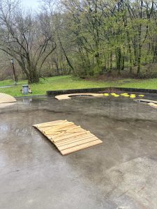 Eine nasse Betonoberfläche im Freien weist eine kleine Holzrampe, gelbe Trittplatten, die einen Pfad bilden, und umgebendes grünes Gras und Bäume mit Blättern an einem bewölkten Tag auf – perfekt für eine MTB- oder Mountainbike-Bewegungseinheit.