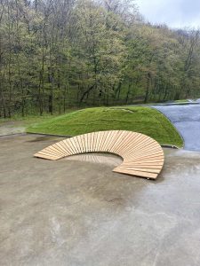 Eine halbrunde Holzbank steht auf einer glatten Betonoberfläche neben einem gewundenen Pfad, mit grasbewachsenen Hügeln und dichten grünen Bäumen im Hintergrund an einem bewölkten Tag.