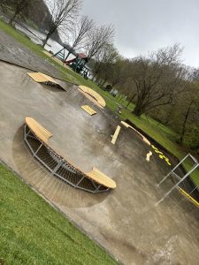 Ein nasser, leerer Skatepark mit Holzbänken an einem bewölkten, regnerischen Tag. Bäume, eine Rasenfläche und im Hintergrund ein überdachter Spielplatz. Der glänzende Boden glänzt wie eine Piste nach dem Regen – perfekt zum Träumen von Mountainbike-Abenteuern.