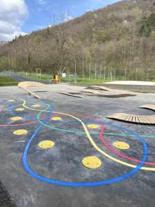 Ein farbenfroher Outdoor-Spielplatz mit gewundenen Gleisen und gelben Kreisen auf dem Boden sowie verstreuten Holzrampen. Eine Person in einer orangefarbenen Weste arbeitet in der Nähe, und im Hintergrund säumen Bäume den Hang – perfekt für Mountainbike-Fans.