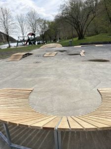 Eine runde Holzbank umgibt eine Betonfläche mit verstreuten Holzrampen und bildet so eine einzigartige Mountainbike-Strecke. Im Hintergrund befinden sich Bäume und ein Spielplatz mit Schaukeln in der Nähe eines Flusses unter einem teilweise bewölkten Himmel.